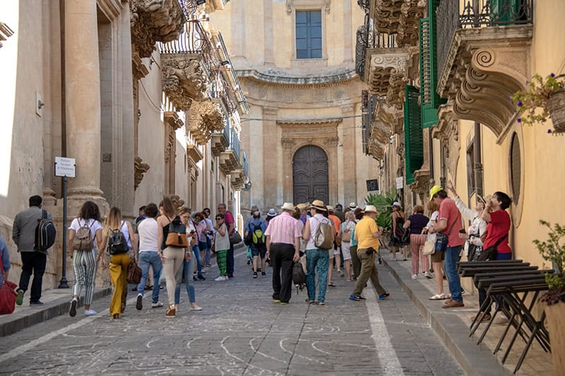people walking along a street with Baroque building while seeing Sicily by car