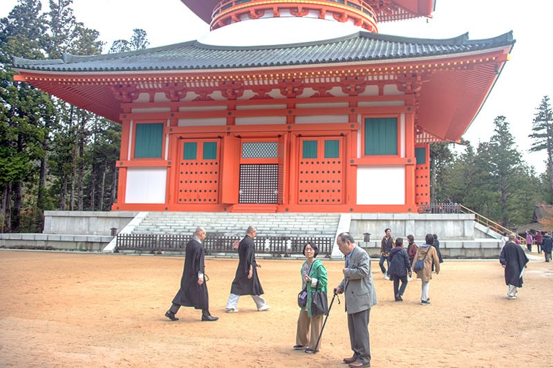 people and monks walking by a vermilion pagoda in Koyasan Japan