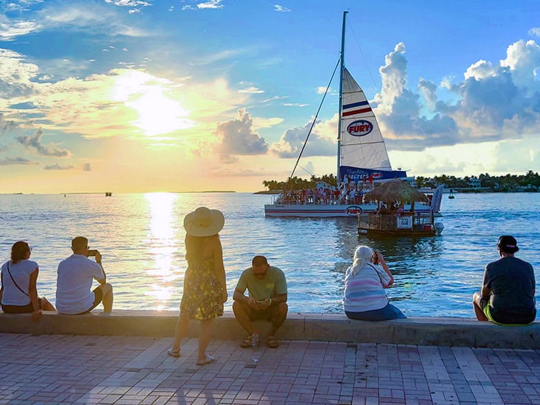 people sitting on a wall watching a sailboat at sunset